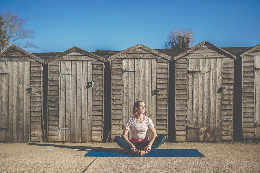 Claire sitting cross-legged on a yoga mat outdoors, smiling and looking up — representing calm movement and nervous system wellbeing